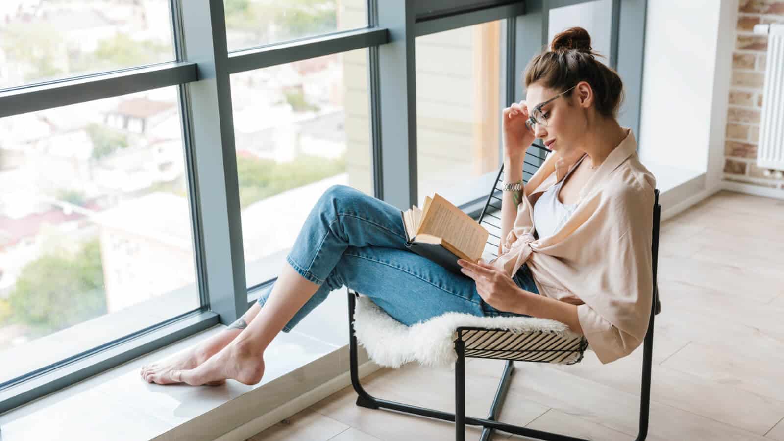 Junge Frau liest Marketing Buch zu Hause auf dem Stuhl.