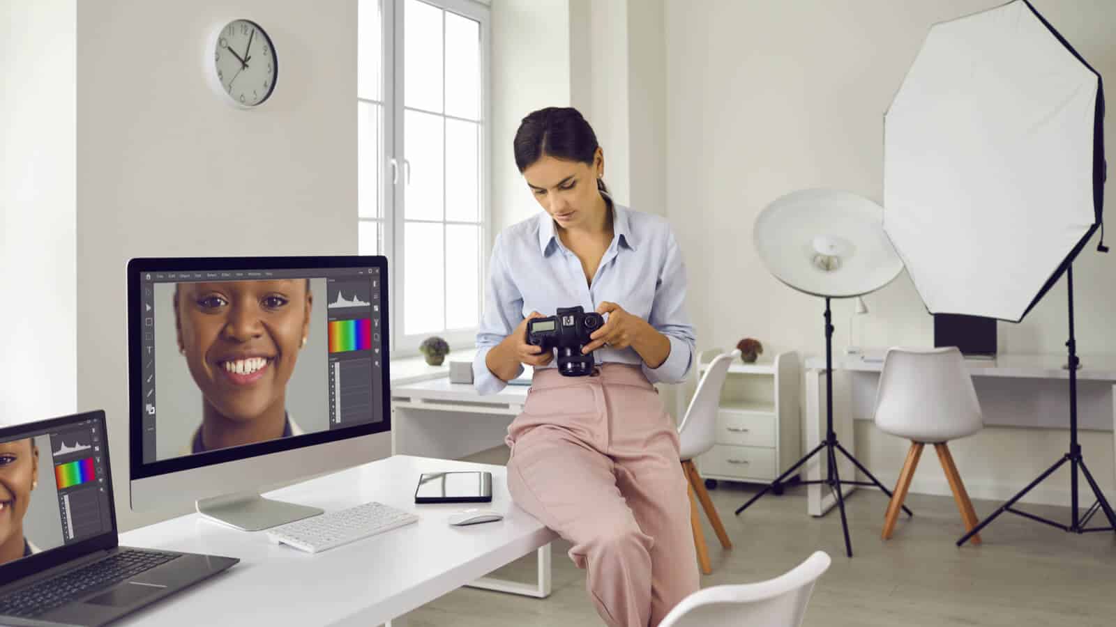 Eine Frau sitzt mit Kamera in der Hand auf dem Tisch und hat ein Bildbearbeitungspogramm offen auf dem Laptop.