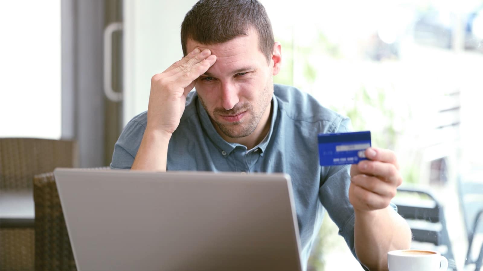 Ein Mann sitzt frustriert am Laptop und bezahlt etwas mit seiner Kreditkarte.