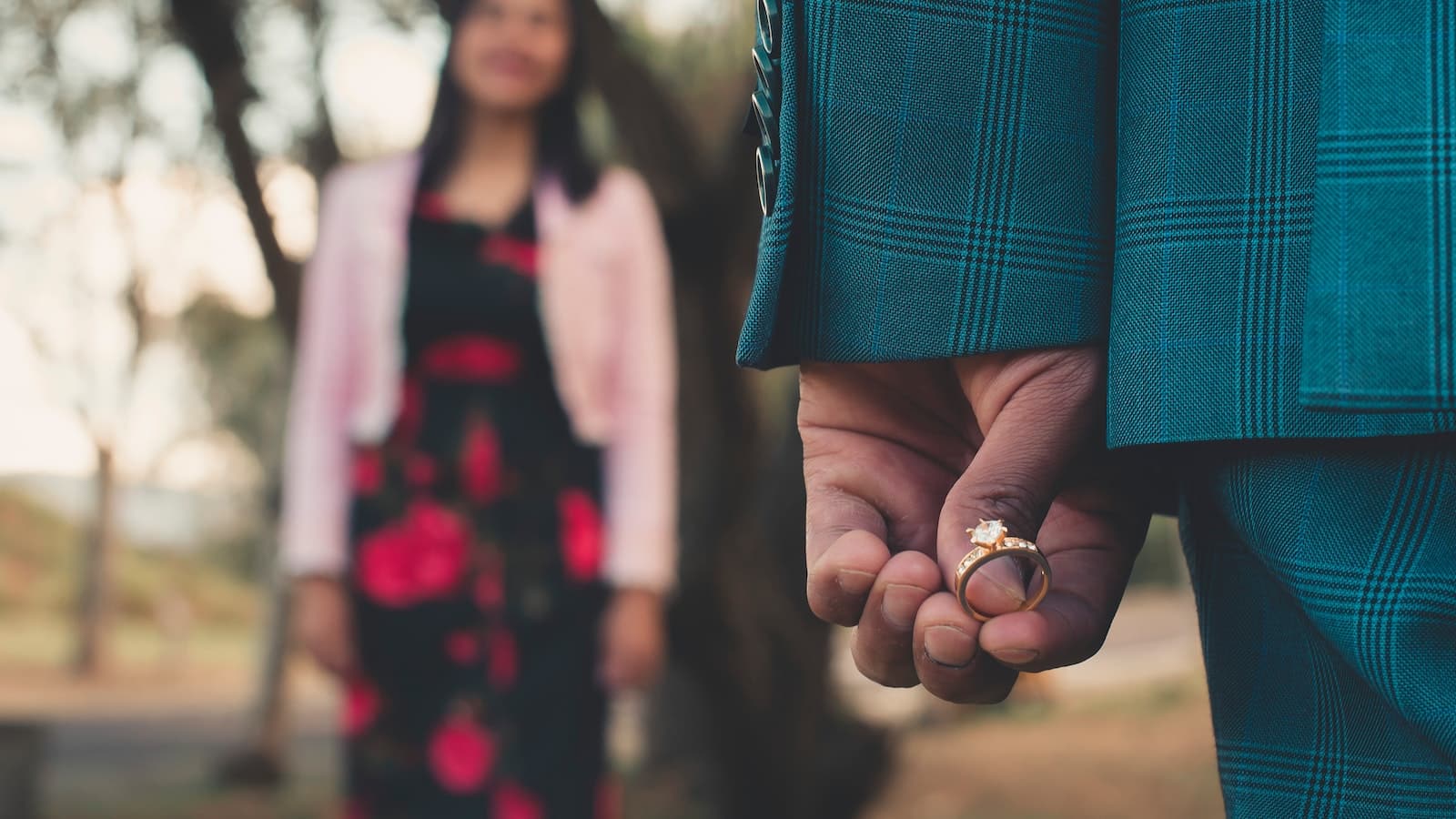 Ein Mann hàlt einen Verlobungsring in der Hand. Im Hintergrund steht eine Frau.