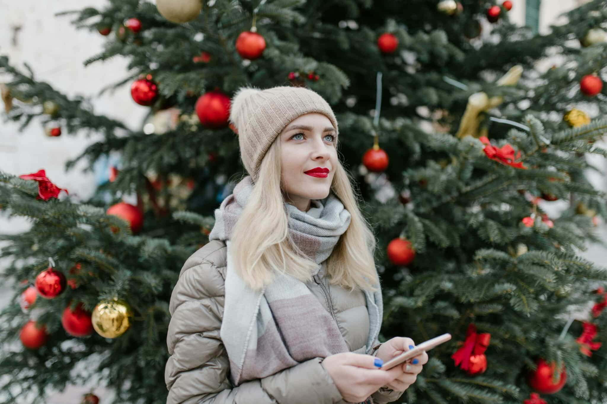 Eine Frau hält ein Smartphone und steht vor einem dekorierten Weihnachtsbaum.