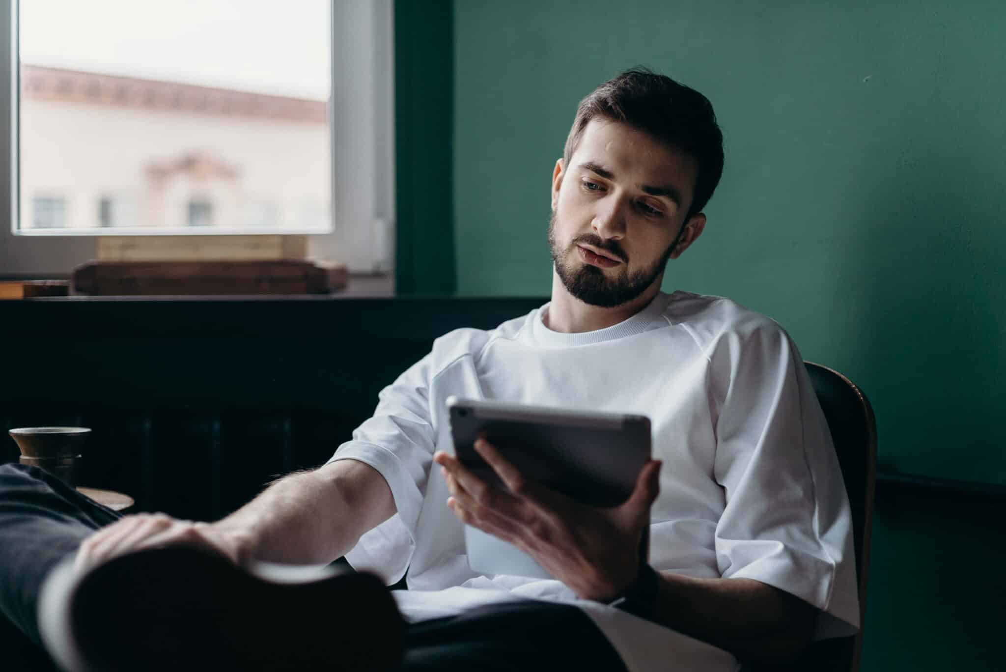 Ein junger Mann im weissen Pullover sitzt entspannt in einem Sessel am Fenster und blickt nachdenklich auf das Tablet in seiner Hand.